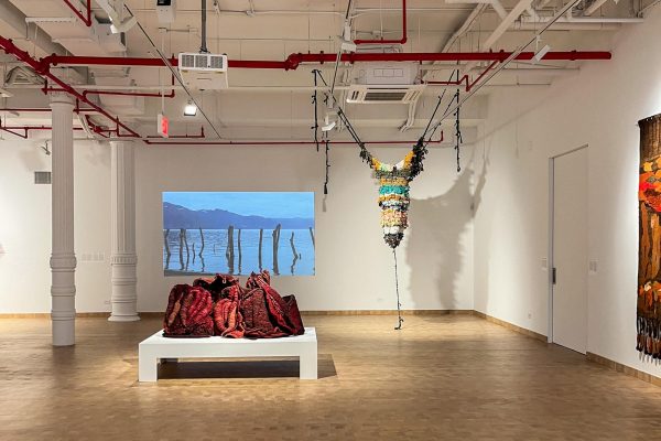 A screen displayed on the wall of wooden sticks in water with a mountain in the background, an art piece displayed dangling from the ceiling and a red art piece displayed on a platform.