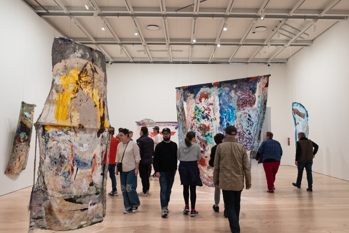 People walk around a large gallery room, looking at multicolored art pieces made of acrylic and metal hanging from the ceiling.