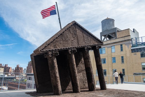 A recreation of the White House facade, made out of earth and dirt, on an outdoor terrace. An upside-down American flag flies above it, and there is an earth floor in front of the sculpture.