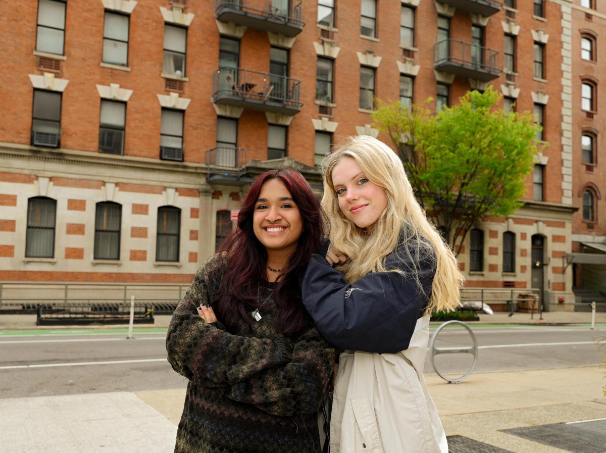 Two people stand on the sidewalk, posing and smiling in front of a row of buildings.