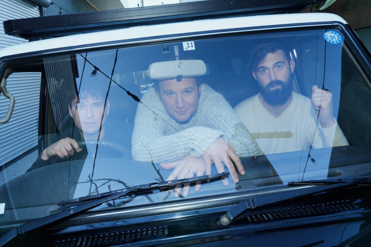 Three members of Vampire Weekend posing in the front seat of a car and looking at the camera through the car's front windshield.