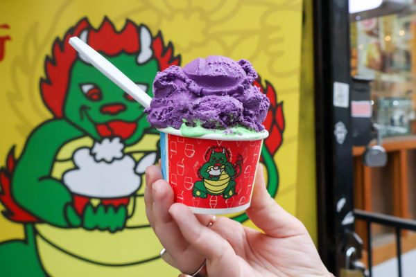 A hand holds a cup of purple and green ice cream in front of a storefront.