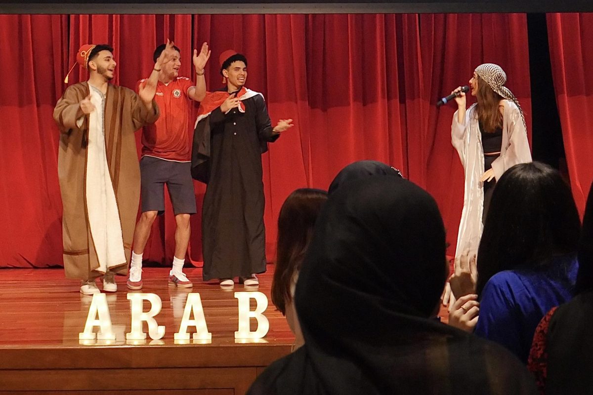 Three men dancing and clapping while a woman sings on a stage. Light up letters are at the front of the stage spelled “ARAB.”