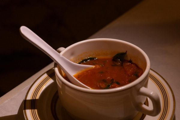 A red-orange broth with green vegetables inside a white bowl with soup soon on a plate. 