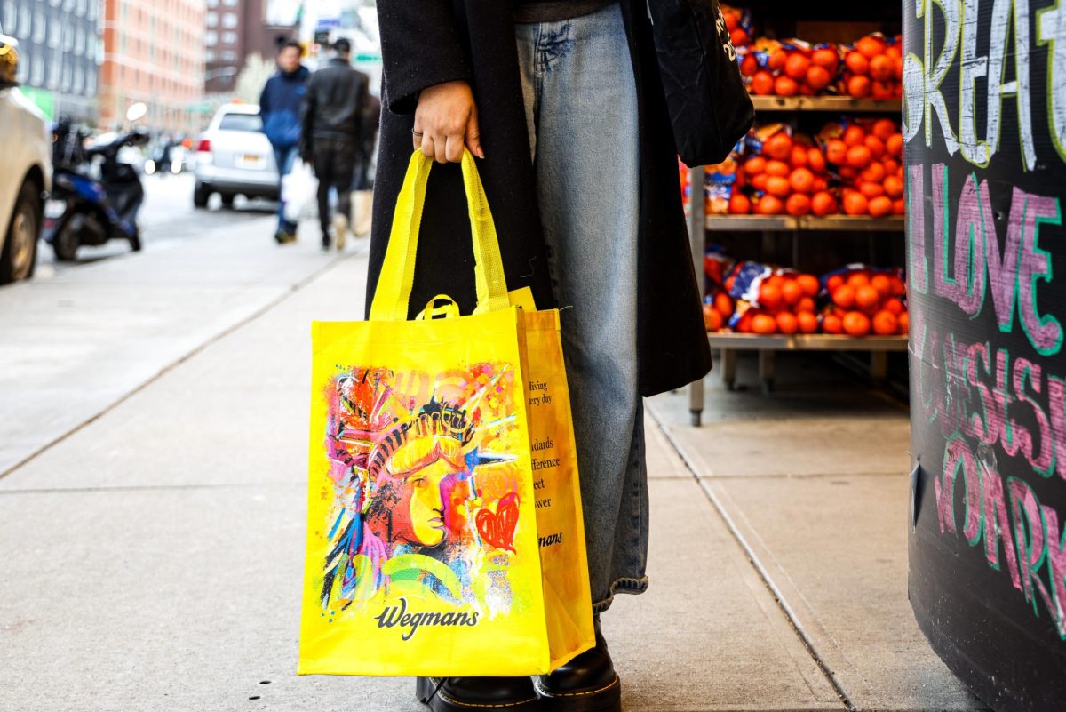 Lower half of a person in front of a market holding a yellow Wegmans tote bag.