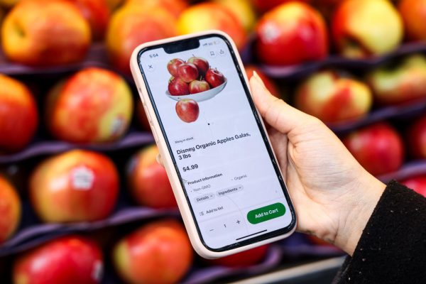 A hand holding a phone with a grocery app open on a listing for apples. There are piles of apples in the background.