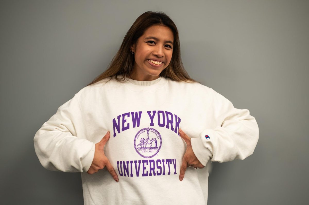 A girl gesturing to her white sweatshirt with the words “NEW YORK UNIVERSITY.” There is a stylized logo on the front of the sweatshirt.
