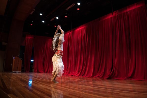A person in white and red clothing dances on a stage with a red curtain.