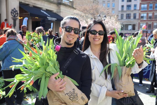 Two people wearing sunglasses stand side by side and smile for the camera while holding paper bags filled with tulips.