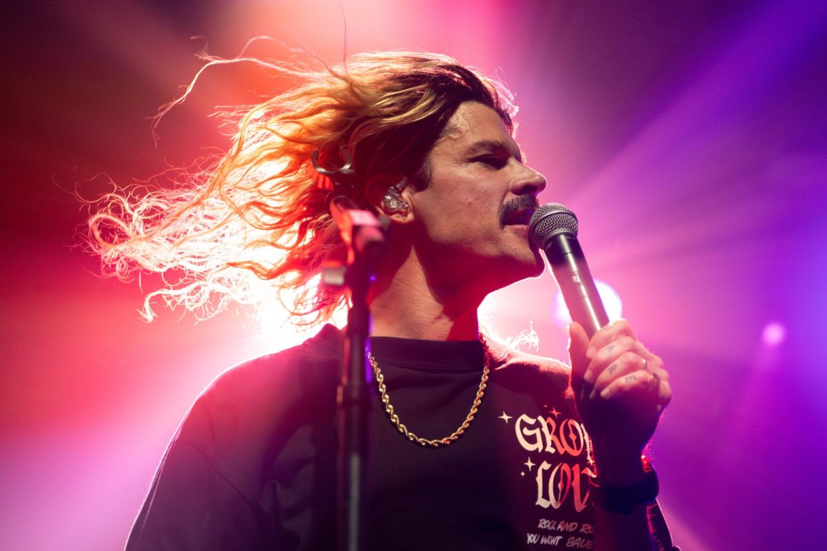 A man with long hair and a mustache sings into a microphone, pink and purple lights outline him.