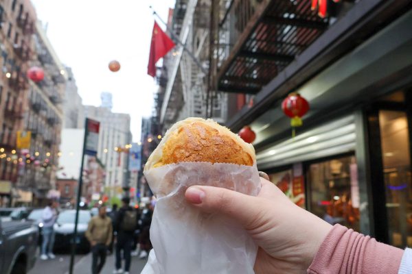 A person’s hand holding a pastry in a white wrapper with a Chinatown street in the background.