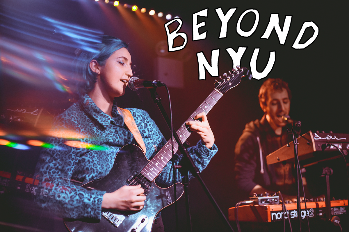 A woman with blue hair and a blue shirt plays a guitar and sings into a microphone. “BEYOND N.Y.U.” is in white font in the top-right corner.