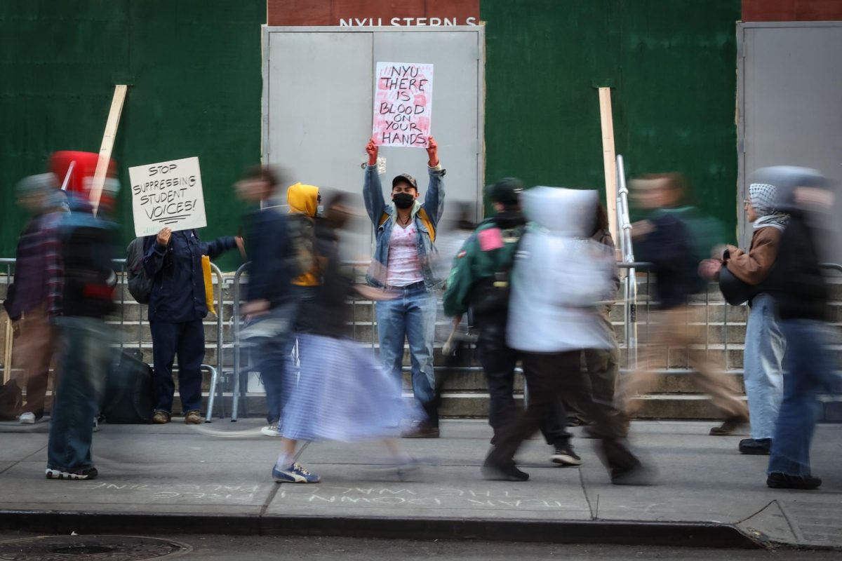 A protester holding a sign that reads “N.Y.U. THERE IS BLOOD ON YOUR HANDS” as other protesters walk in front of them.