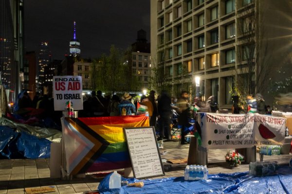 Protesters at an encampment set up in an outdoor place at night. In front of them are signs, three small Palestinian flags and a large pride flag.