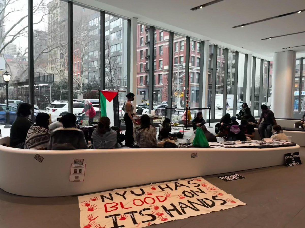 Students gathered in the Paulson Center lobby with a sign saying “N.Y.U. HAS BLOOD ON ITS HANDS” in black and red font with red paint handprints. A Palestinian flag is hanging on the window behind them.