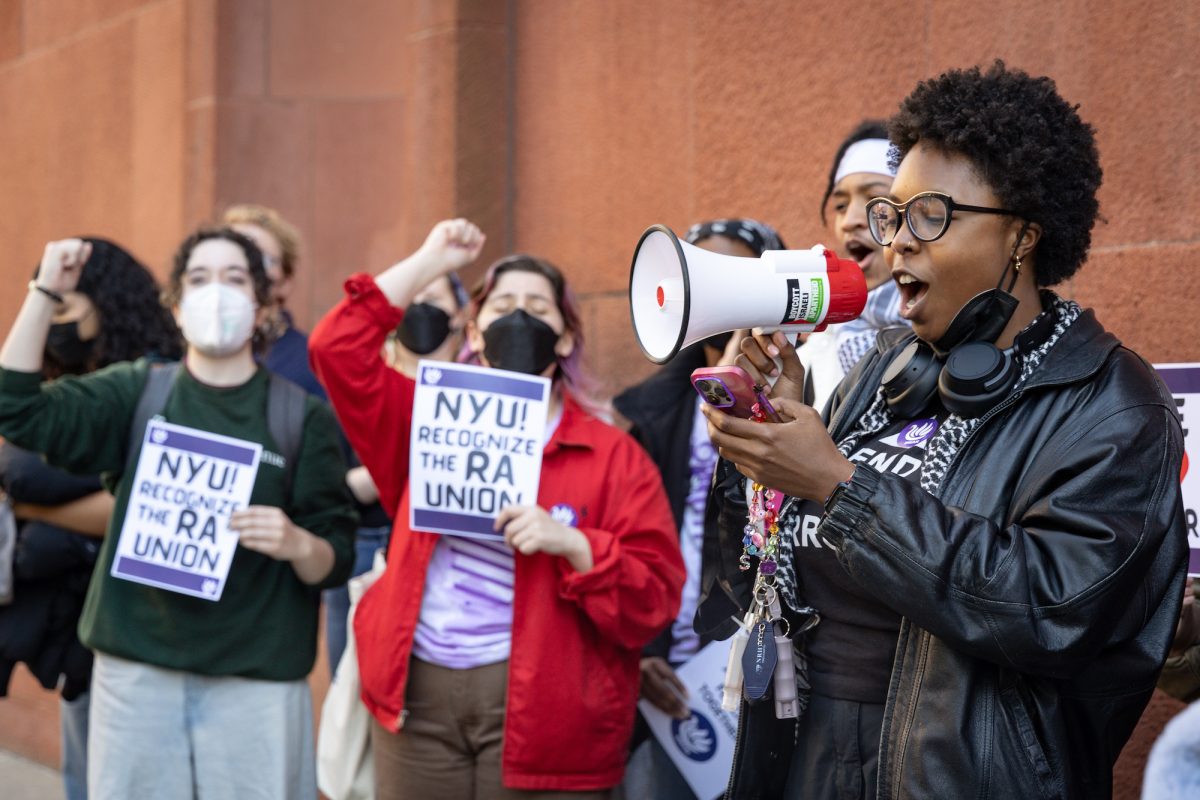 A person shouts into a megaphone. Behind them, people hold flyers that say “NYU! RECOGNIZE THE RA UNION” with one hand and hold their other fists up in the air.