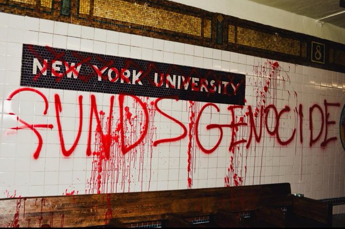Subway station with New York University signage covered in red spray paint splatters and the tag “FUNDS GENOCIDE.”