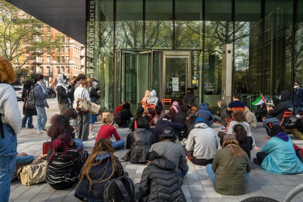 A group of people sit on the ground outside of a glass building.