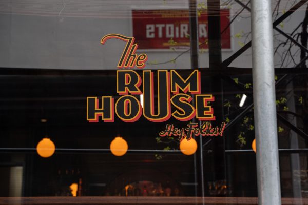 Window of a bar with the sign that says “THE RUM HOUSE: HEY FOLKS!” in black, orange and yellow calligraphy font. 
