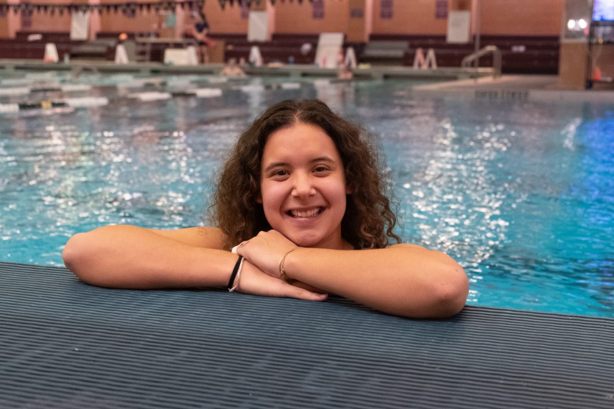 A girl with brown hair sitting in a pool crosses her arm on the pool’s ledge.