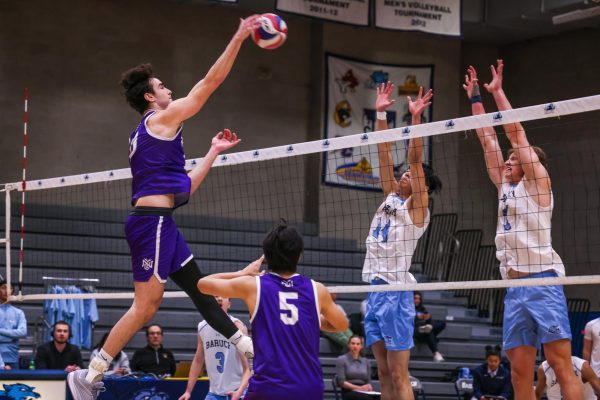 A man in a purple uniform smashes a volleyball over a net. On the other side, two defenders in white uniforms jump up to block him.