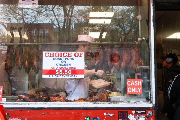 A man in the back of a restaurant with a line of pork shown in the front, and the words “CHOICE OF ROAST PORK OR CHICKEN ON A SMALL RICE $5.50 ALL DAY EVERYDAY,” displayed on the front of the restaurant.