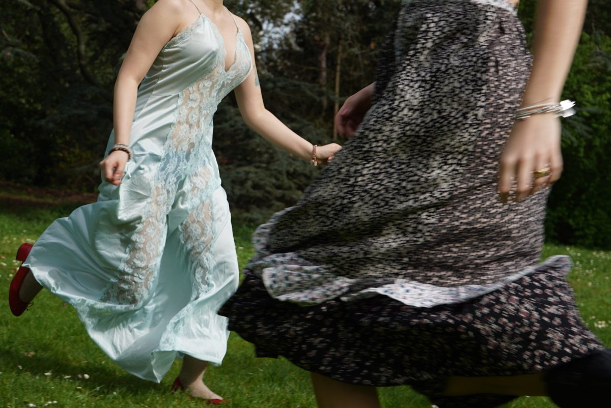 Two people, one in a dress and the other in a skirt, in a photo that cuts off below their heads. The fabric of their clothing is moving in the wind as they move in a grassy field.