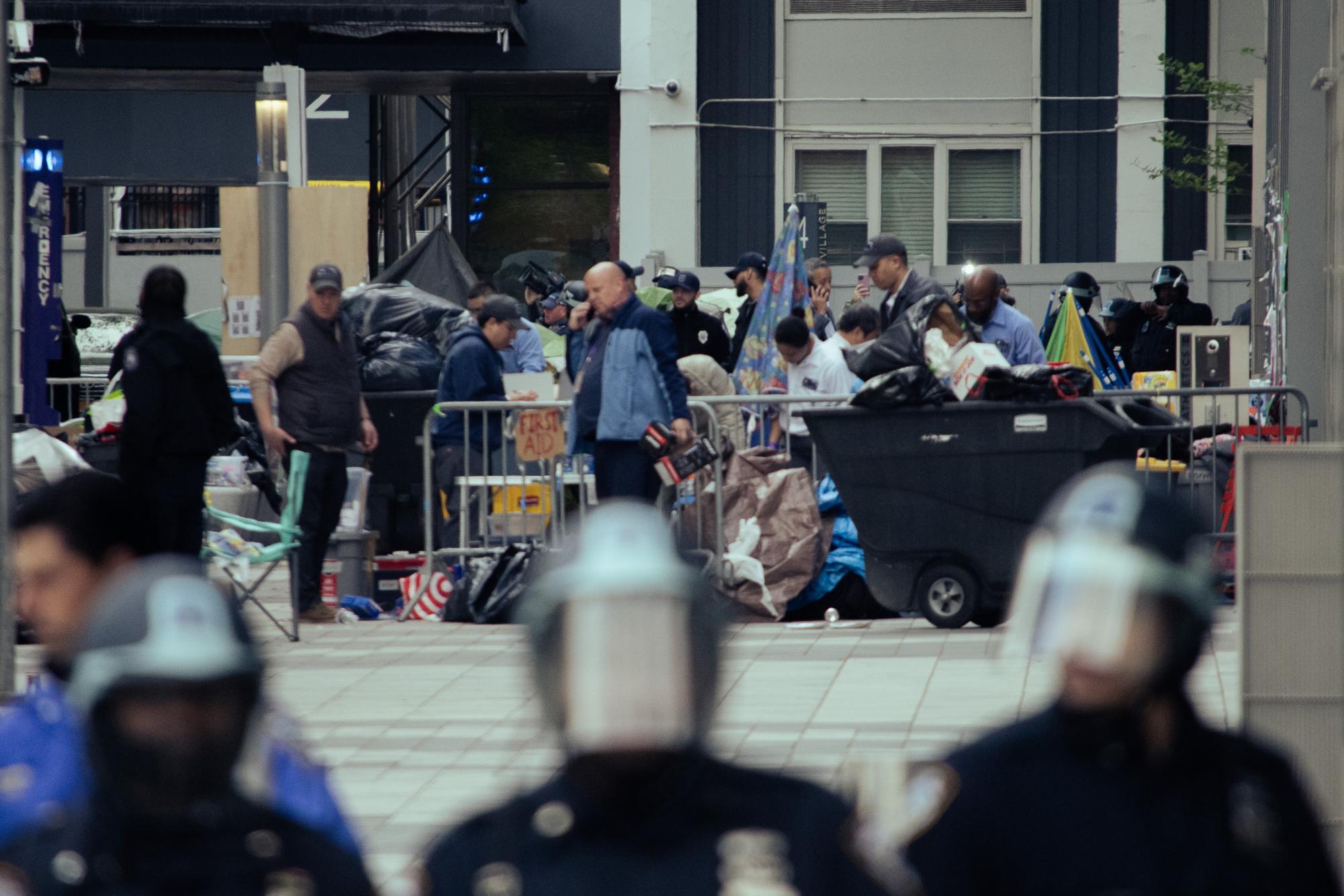 Three N.Y.P.D. officers in riot gear stand in the foreground. Cleaning staff clear out the encampment in the midground and media personnel set up cameras at the back.