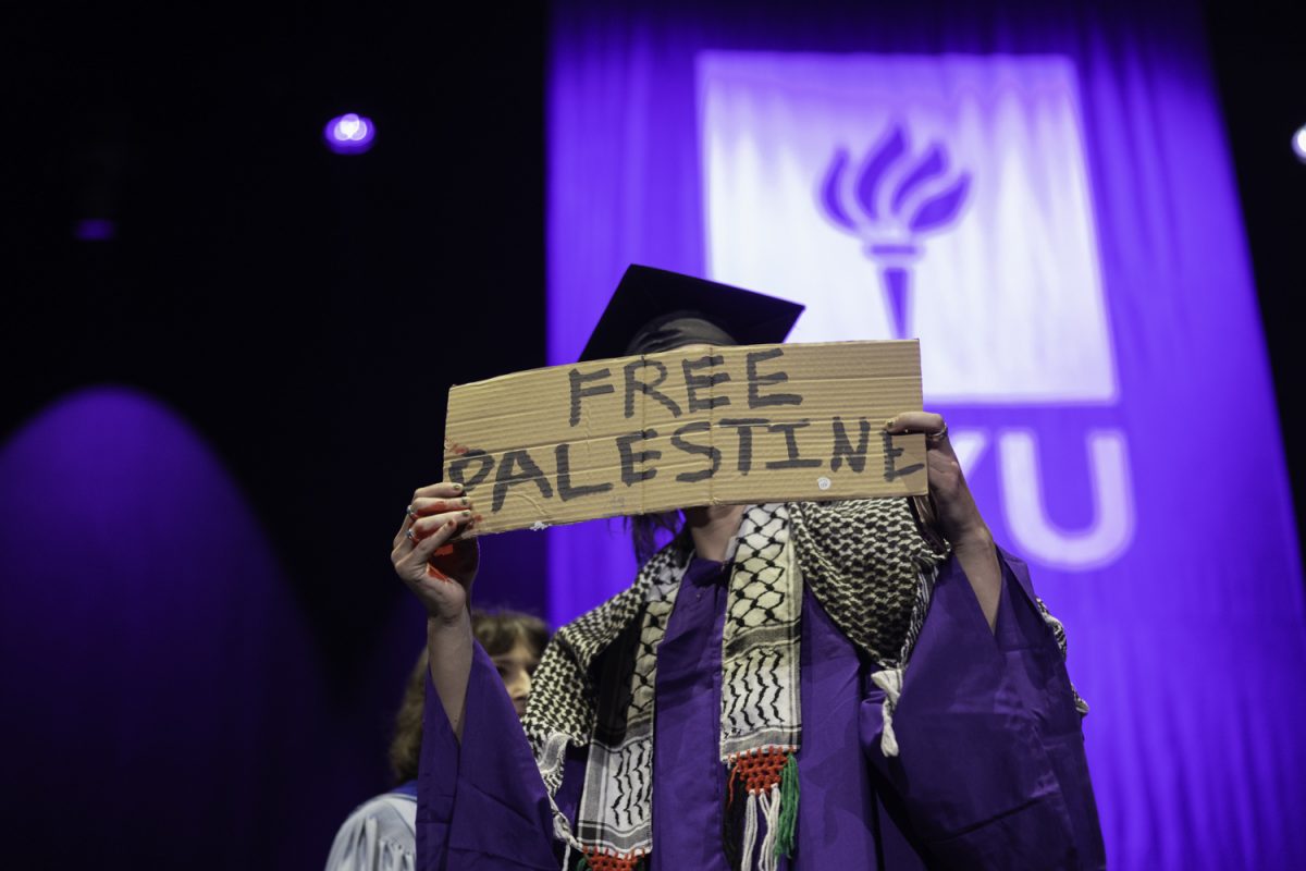 A man in a black mortarboard and purple robe with a white and black scarf holds up a cardboard sign reading “Free Palestine”