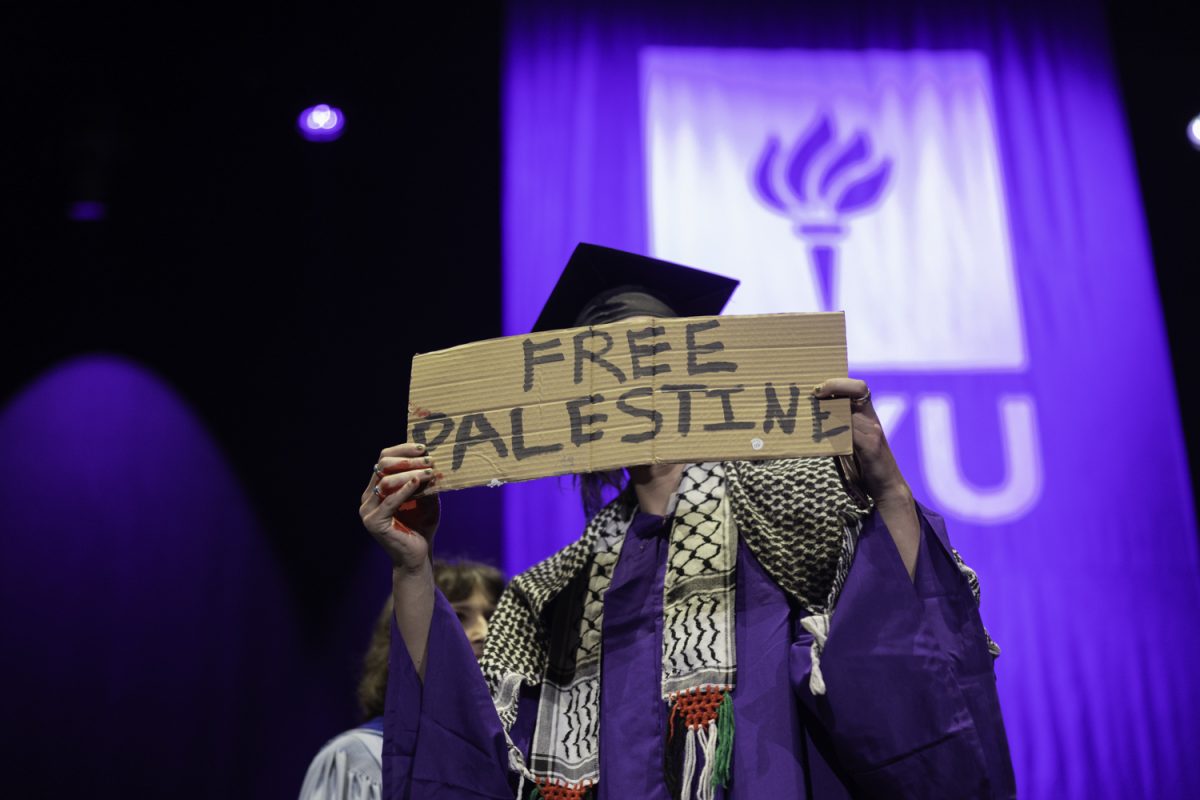 A+man+in+a+black+mortarboard+and+purple+robe+with+a+white+and+black+scarf+holds+up+a+cardboard+sign+reading+%E2%80%9CFree+Palestine%E2%80%9D