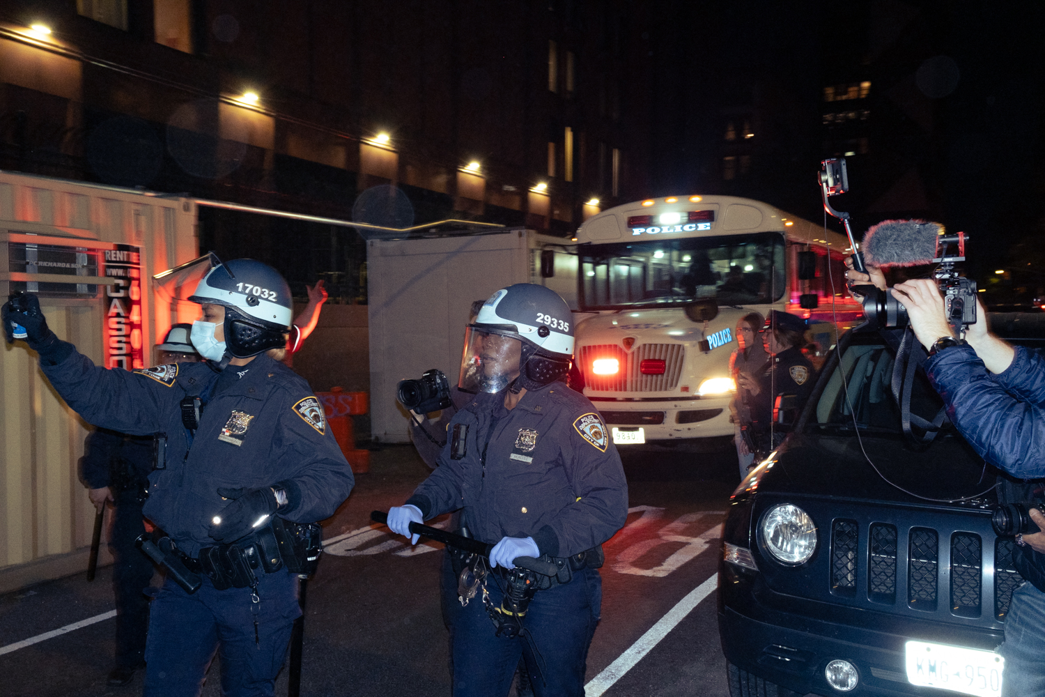 Two N.Y.P.D. officers walk in front of a correctional bus holding pepper spray and batons.
