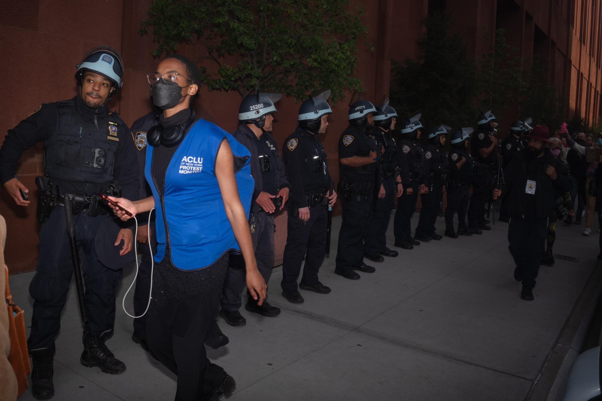 An A.C.LU. legal observer run alongside a line of police in riot gear outside Bobst Library.