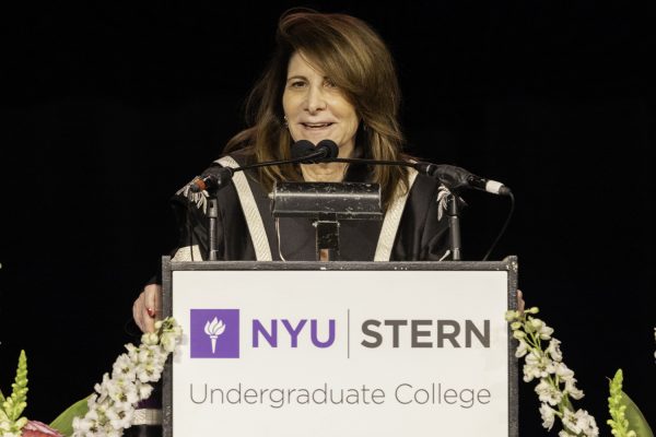 A woman with brown hair and a black robe with white accents stands in front of a podium with a white sign that reads “N.Y.U Stern Undergraduate College”.