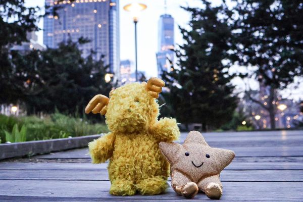 Two stuffed animals, one yellow moose and a beige star, sitting on the ground with buildings in the background.