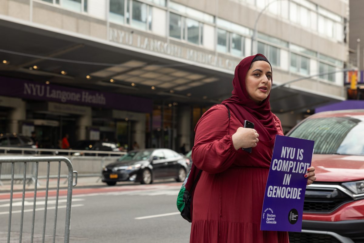 A woman dressed in red and holding a purple sign that reads NYU is Complicit in Genocide.