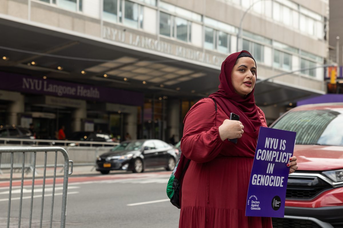 A woman dressed in red and holding a purple sign that reads "NYU is Complicit in Genocide".