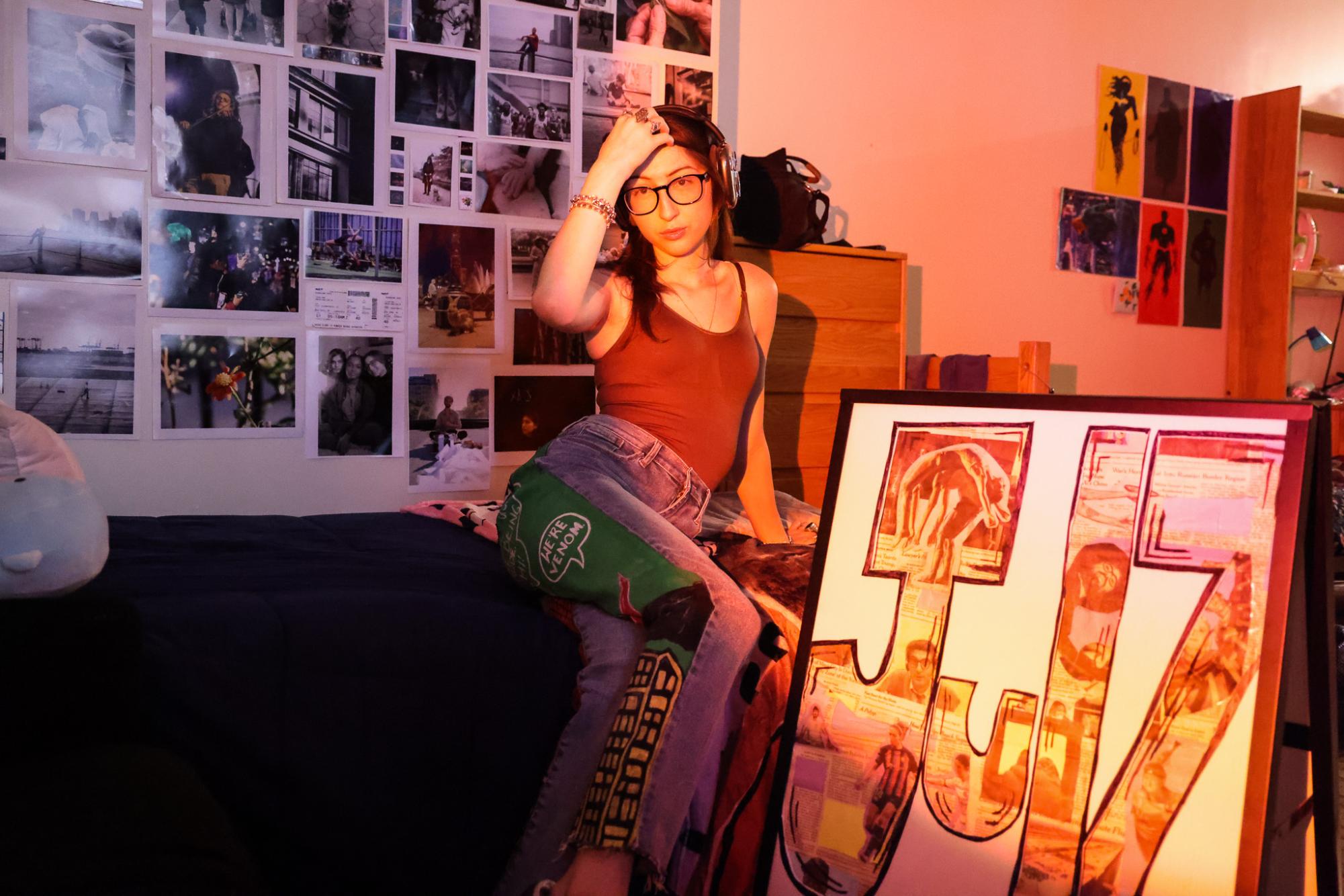 A woman sitting on her bed, posing to show the leg of her painted jeans.