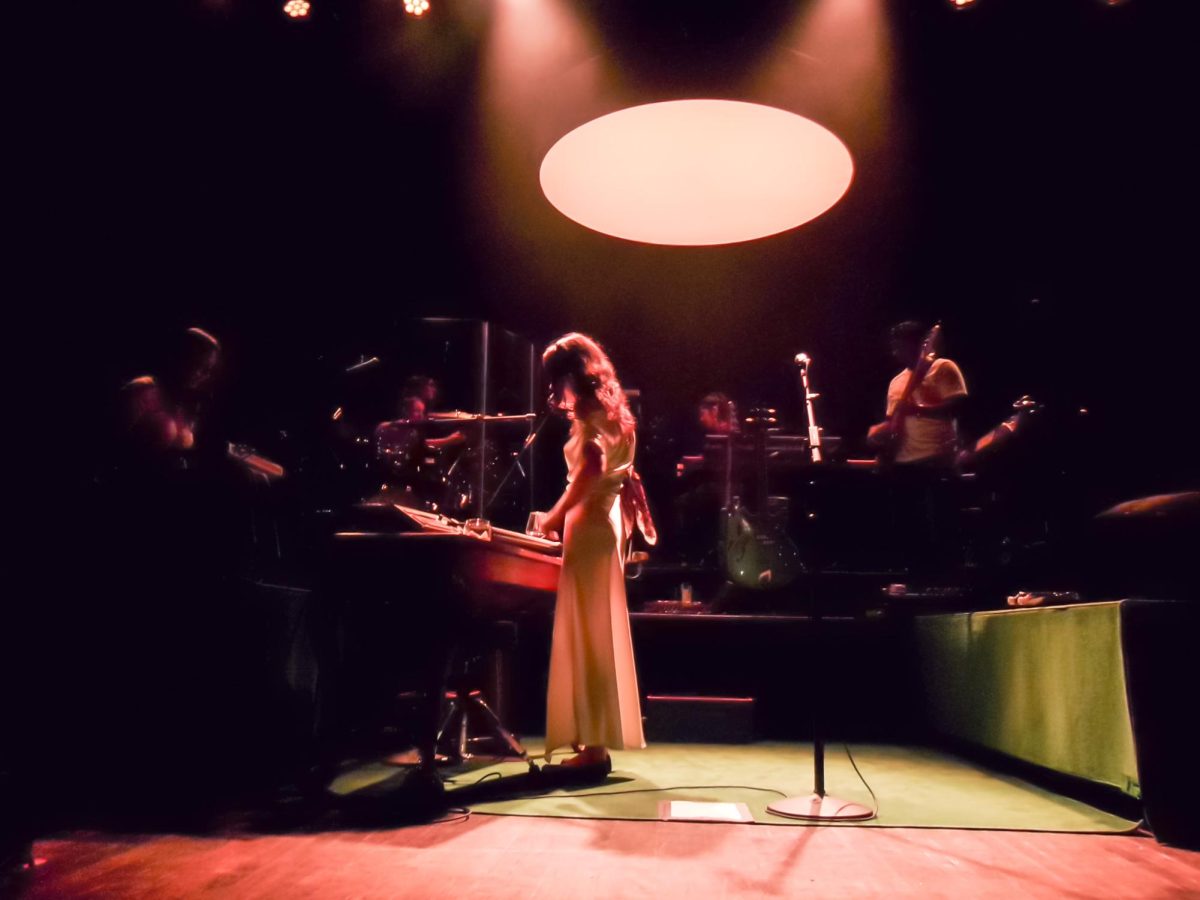 A woman stands playing a keyboard in a long white dress with a band playing guitars and drums on risers behind her.