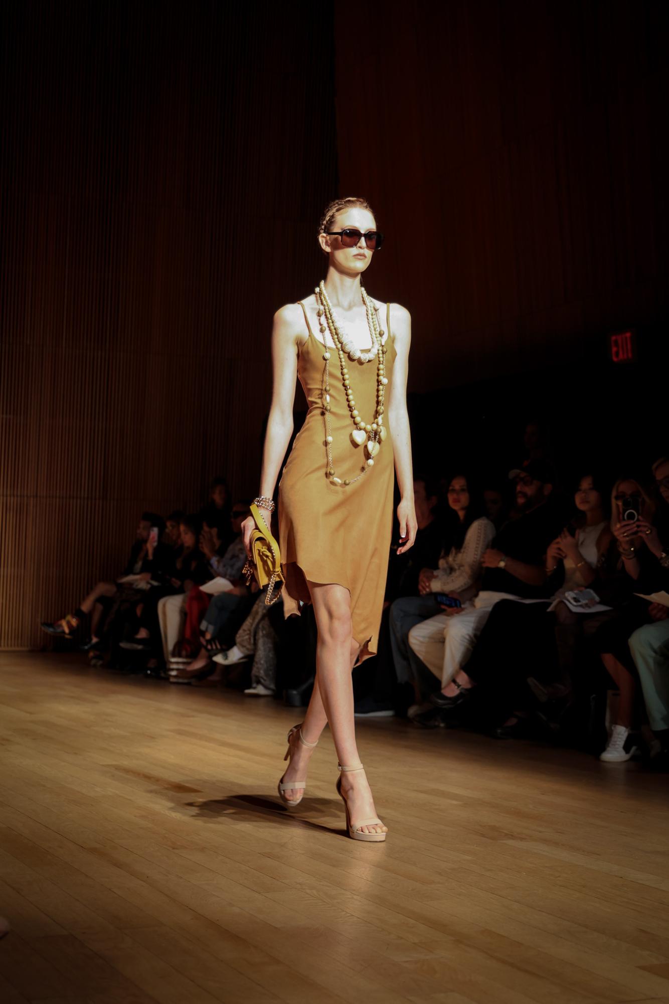 A model in a knee-length, mustard dress with a beaded necklace, chunky sunglasses and a yellow handbag.