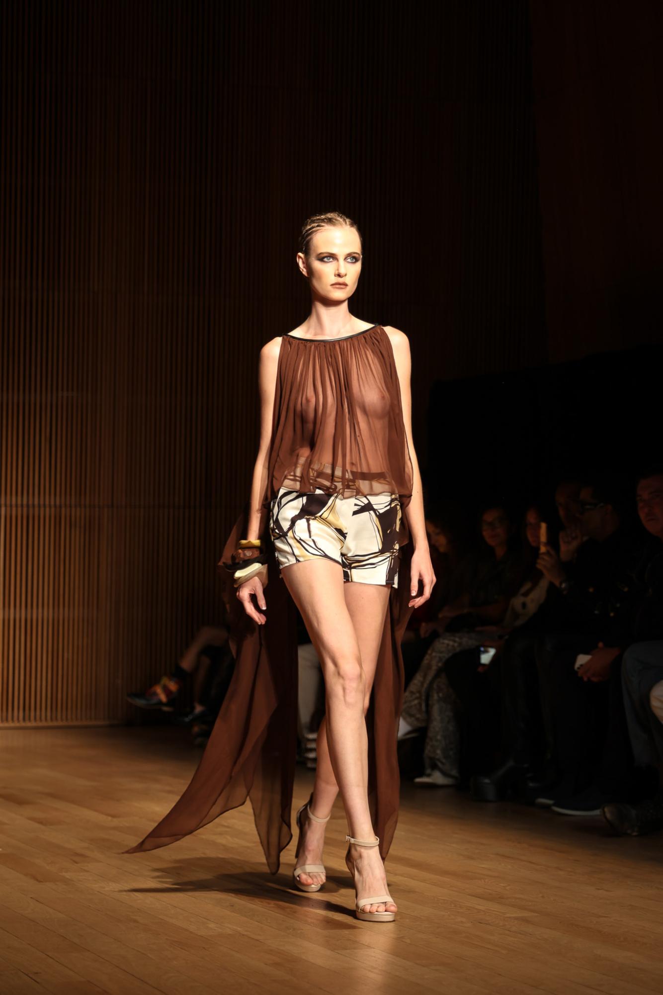 A model wearing a brown, flowy see-through shirt with black, white and yellow shorts and nude heels.