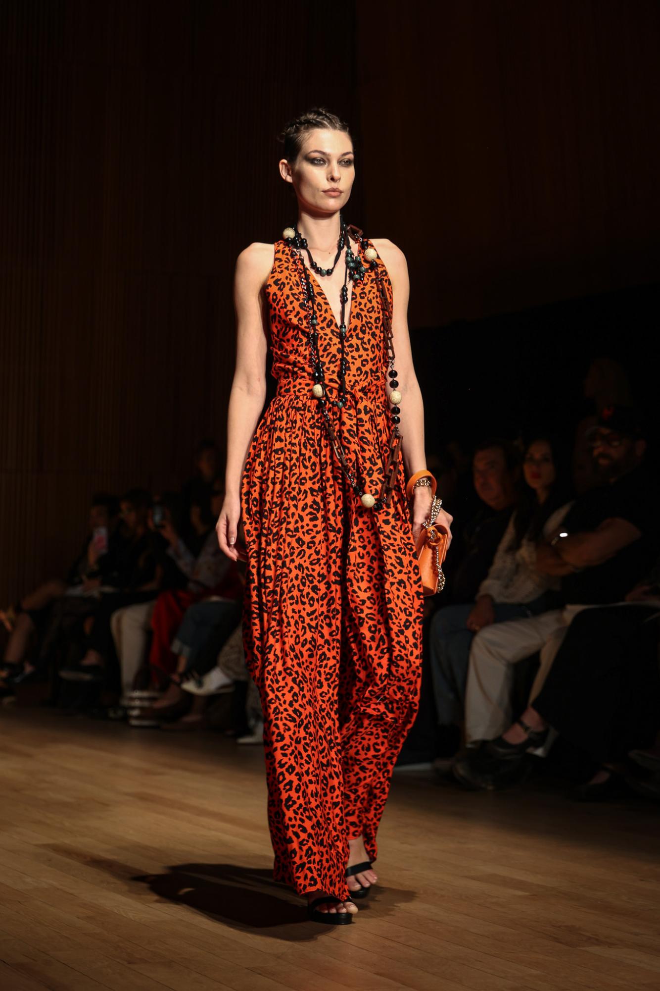 A model in a red, cheetah print pant suit, with a long, black and white beaded necklace. She carries an orange purse in her hand.