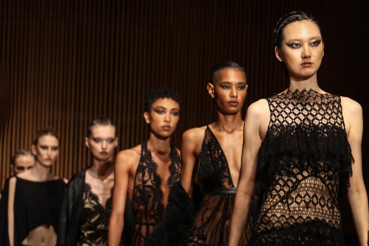 Six models wearing all black, walking down a runway in a single-file line.