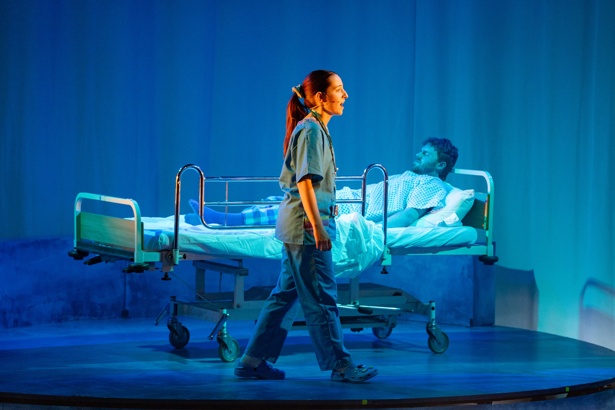 A woman in teal scrubs walks across the stage as a man in a hospital gown lies in the background on a hospital bed.