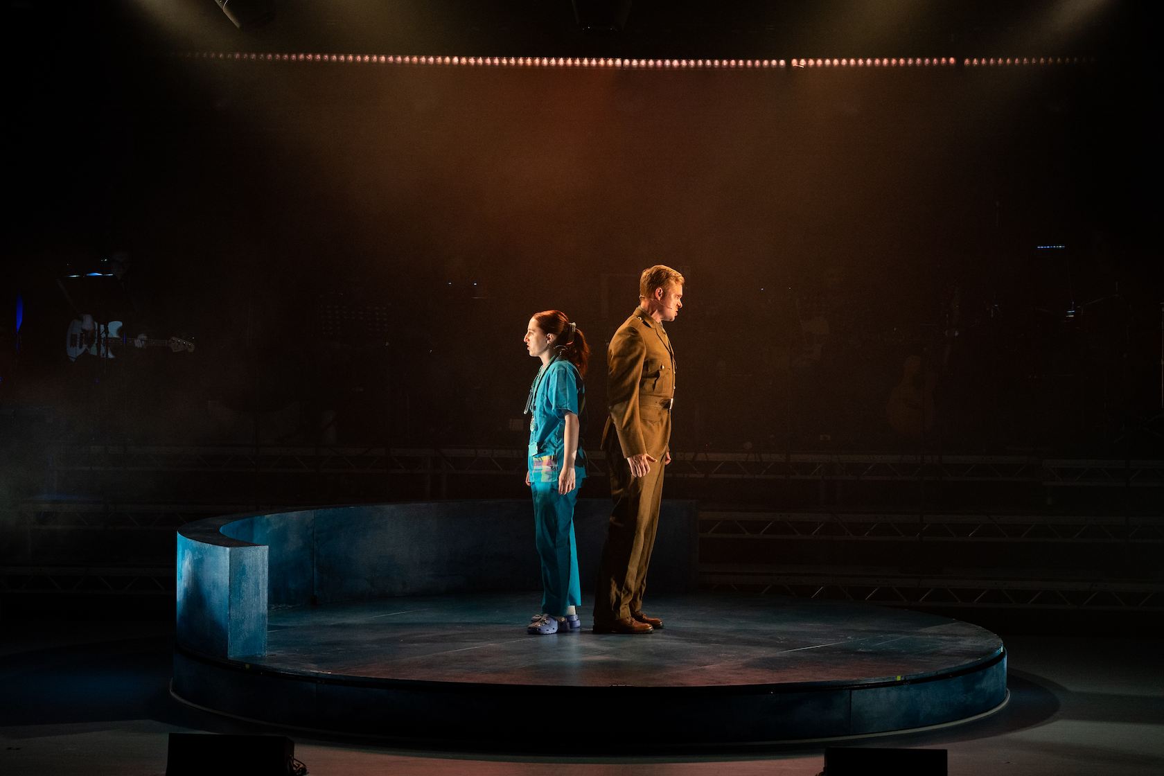 Two people stand back-to-back in the middle of a dark stage. On the left is a woman in scrubs, while a man on the right is wearing a brown military suit.