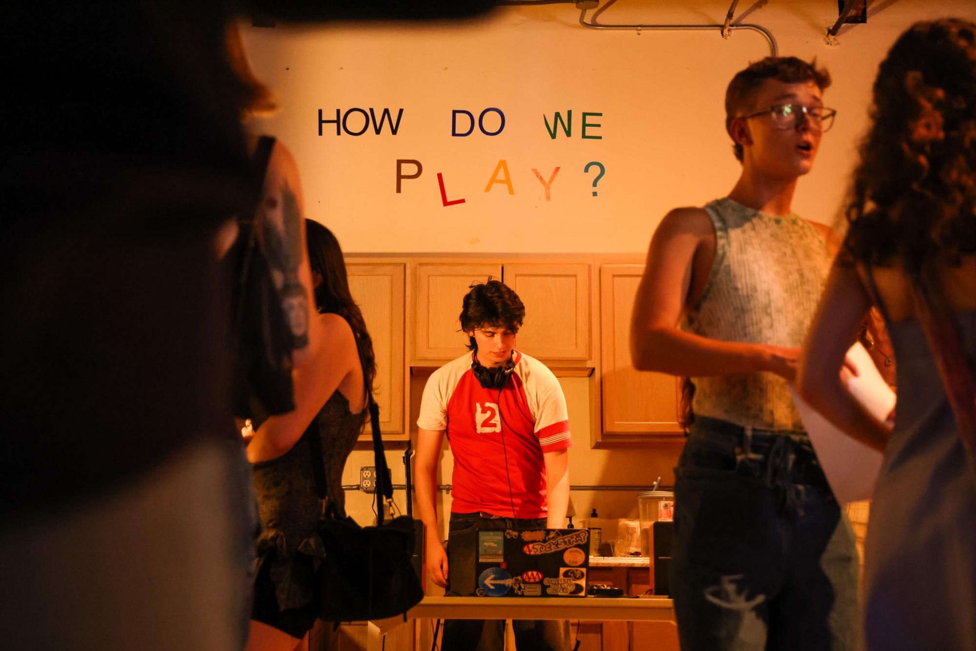 Surrounded by other people, a person in a red shirt stands under letters on a white wall which reads, “HOW DO WE PLAY?” in rainbow colors.