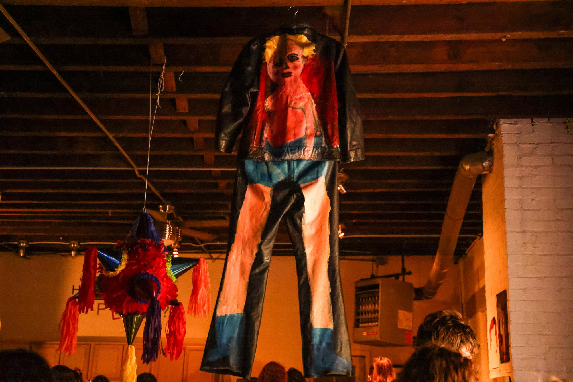 A painted jean jacket and matching pants hang from the ceiling with a piñata hanging behind it.