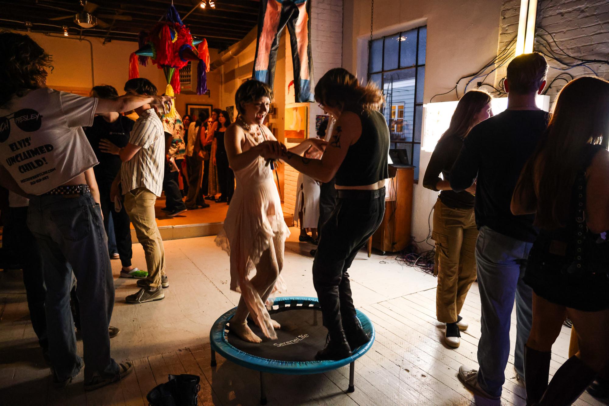 Two people stand in the middle of a dimly-lit room surrounded by others as they dance hand in hand.