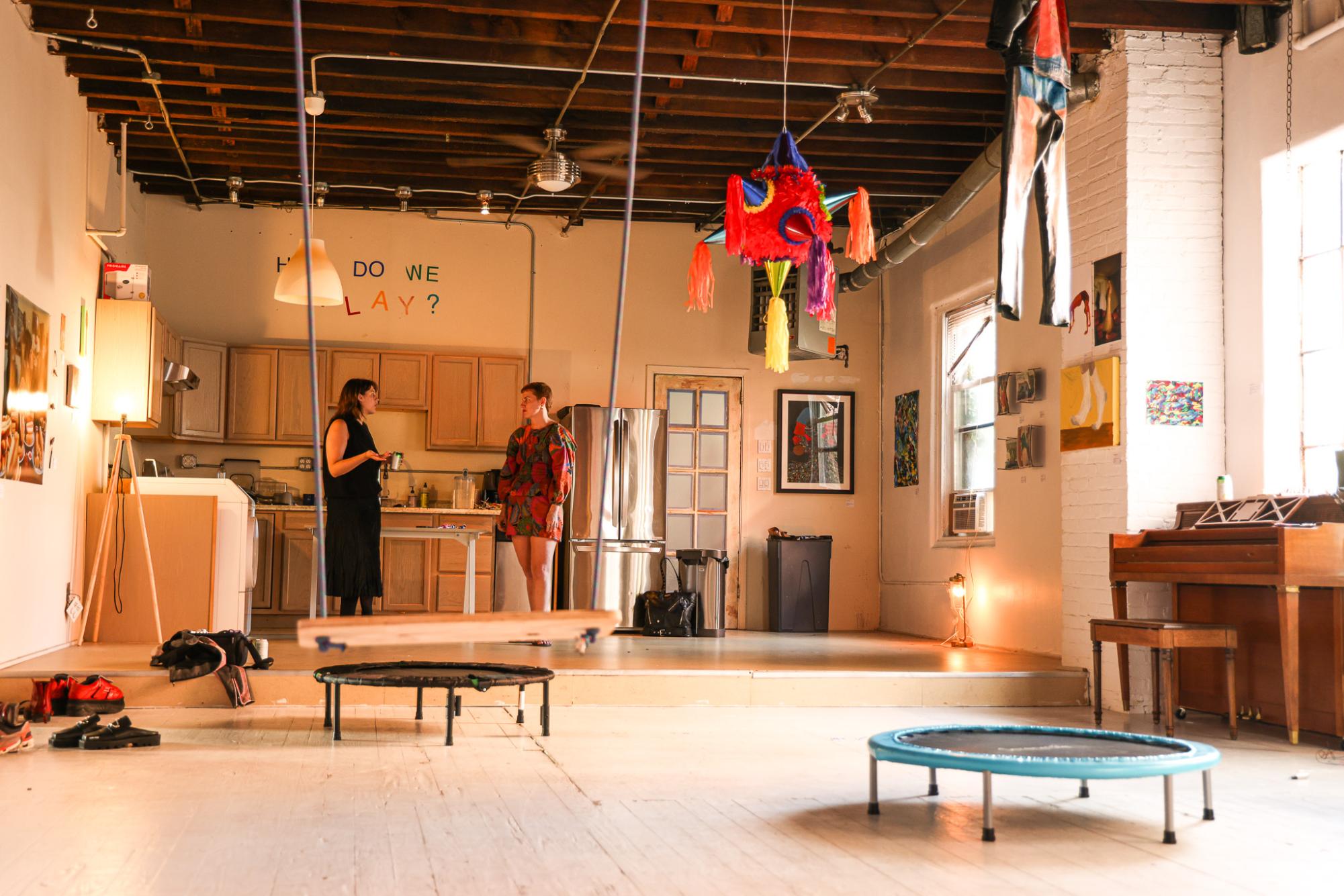 Two people talk while standing in a kitchen with a piñata and jumpsuit hanging from the ceiling and a piano.