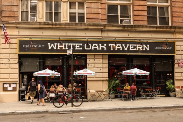 A storefront with the words “WHITE OAK TAVERN” with wooden tables and chairs and mounted umbrellas outfront.