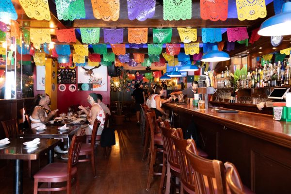 An interior with colorful papel picado hanging above a bar and wooden tables and chairs.